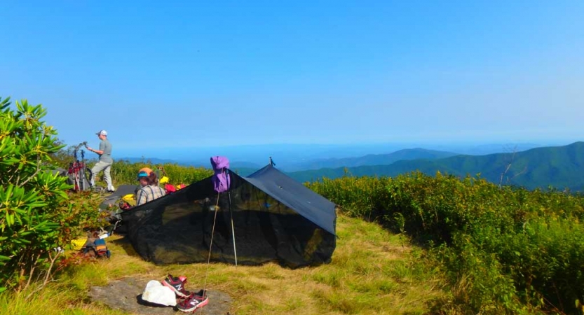 A campsite with a tarp shelter rests high above a green mountain landscape. 